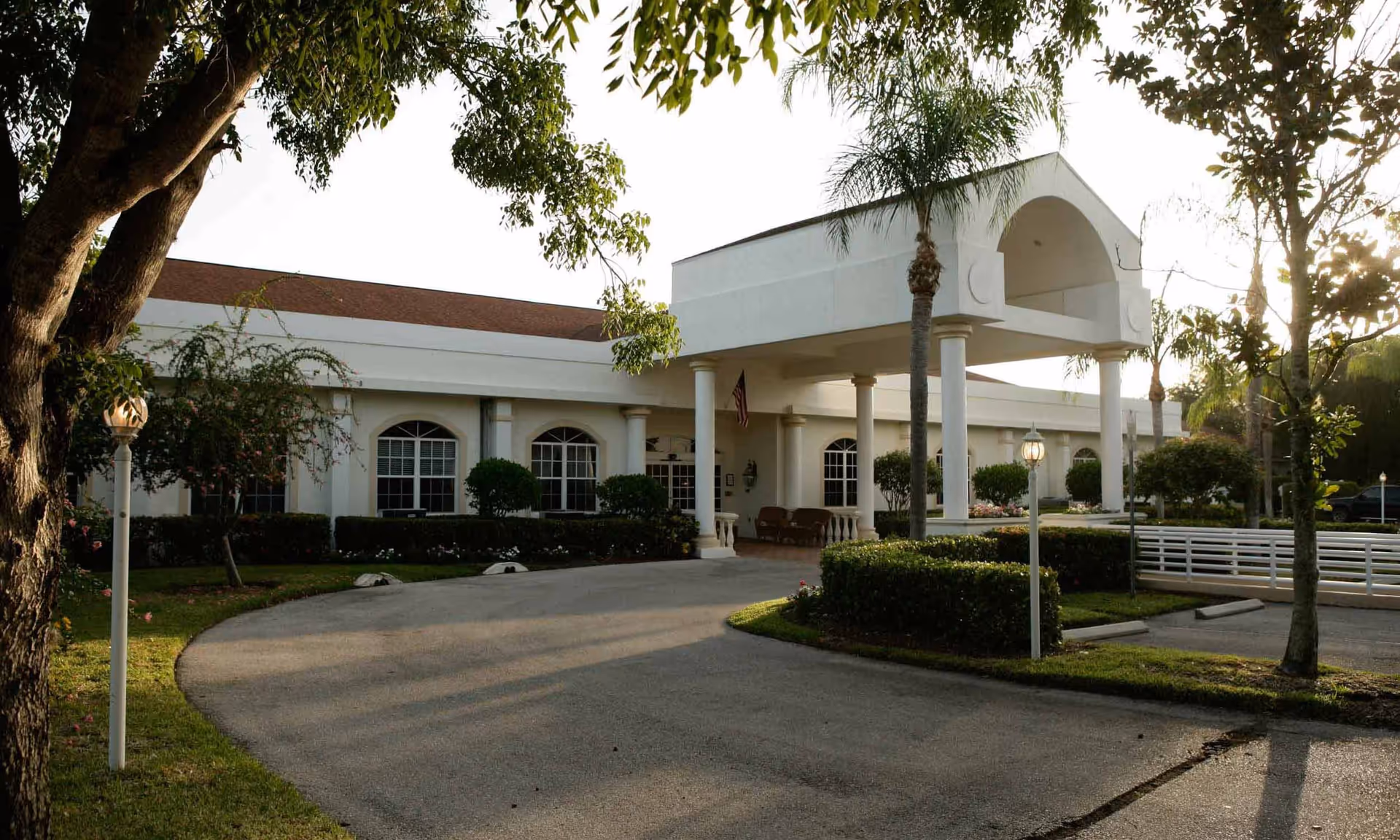 Exterior view of Naples Green Village facility showing a white building with a covered entrance supported by columns, surrounded by trees, bushes, and a driveway.