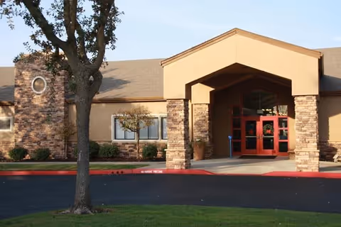 Front entrance of a single-story senior living building with a covered portico, stone columns, red double doors, and a tree in the foreground.