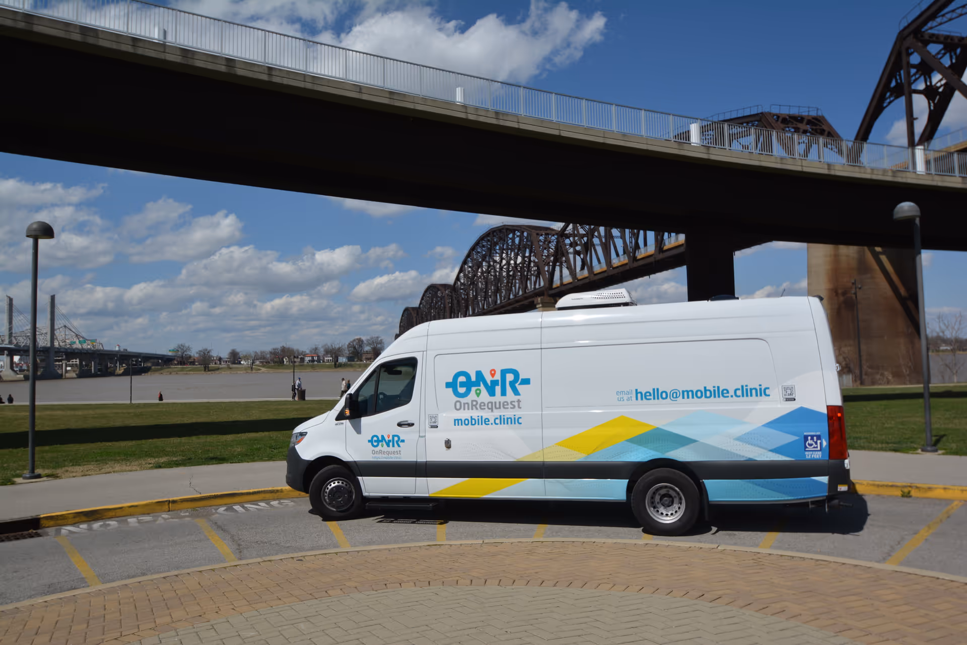 White mobile clinic van with OnRequest branding parked near a river under a bridge.