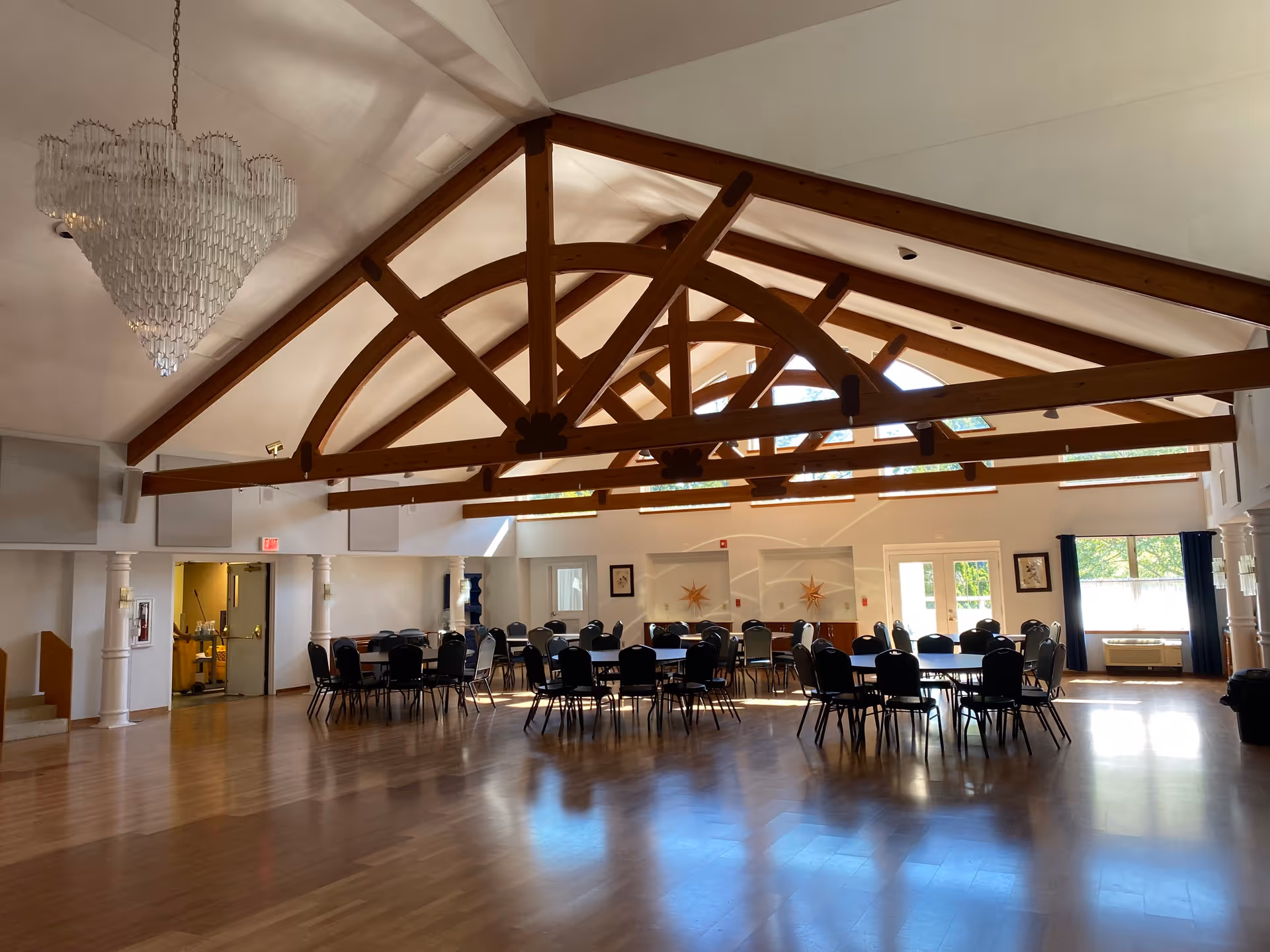 Spacious dining room with wooden floors, multiple round tables with chairs, large wooden ceiling beams, a chandelier, and windows letting in natural light.