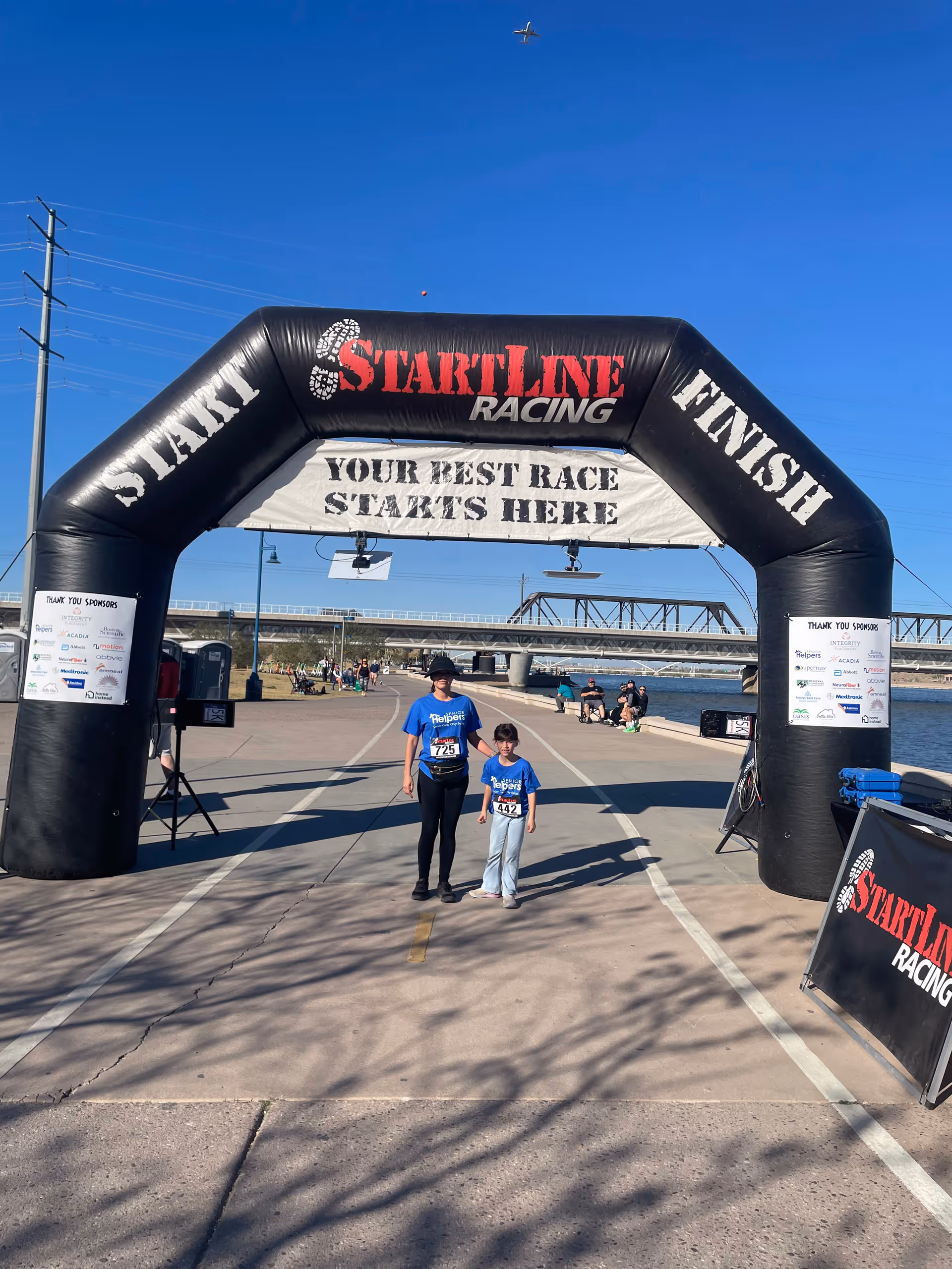 Two people pose under a Start Line Racing arch on an outdoor waterfront path.