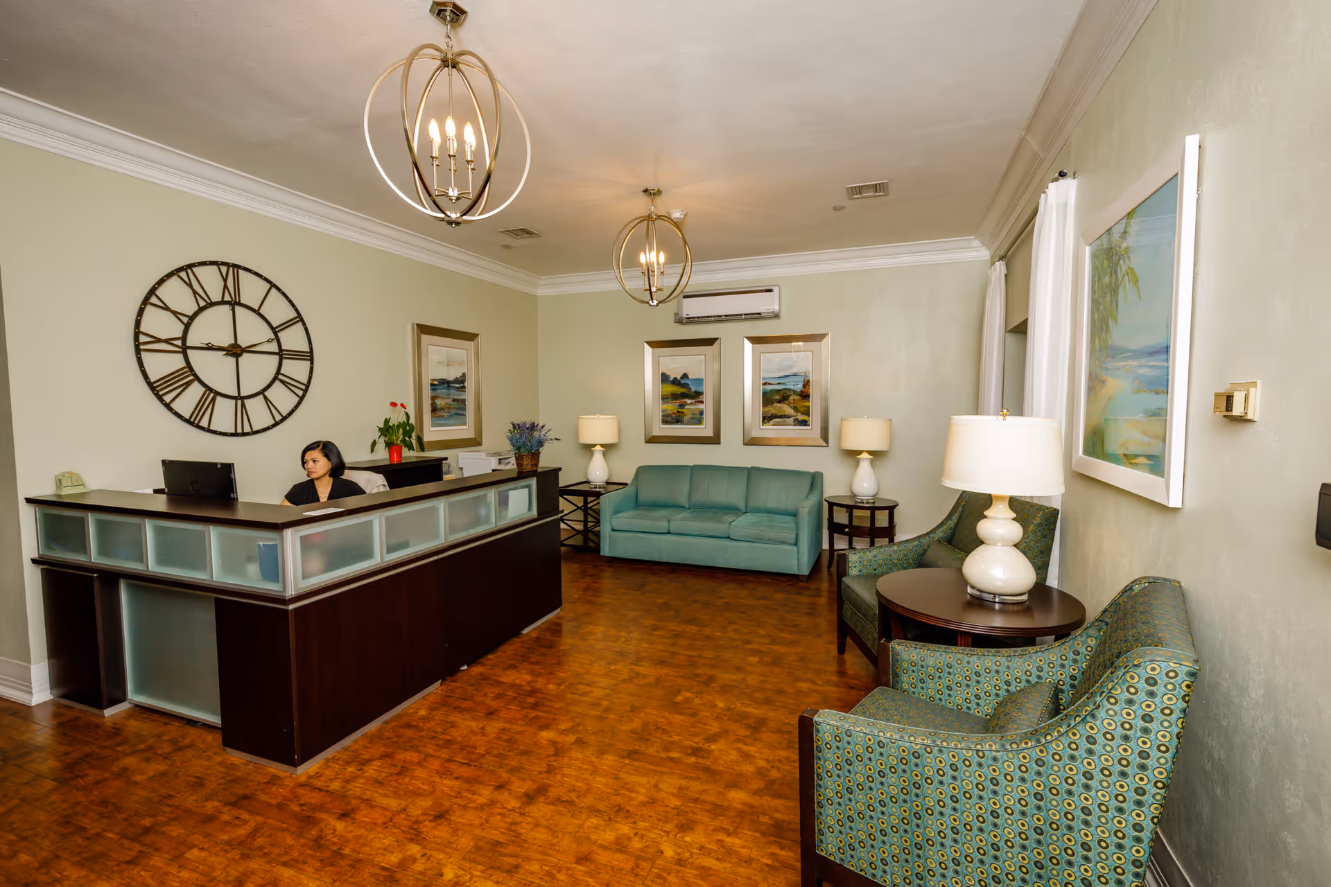Reception desk with a staff member in a furnished waiting area featuring a teal sofa, patterned chairs, lamps, wall art, and a large decorative clock.