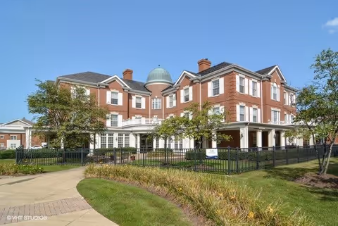 Red-brick three-story senior living building with a small dome, wraparound porch, fenced lawn and walkway under a clear blue sky.