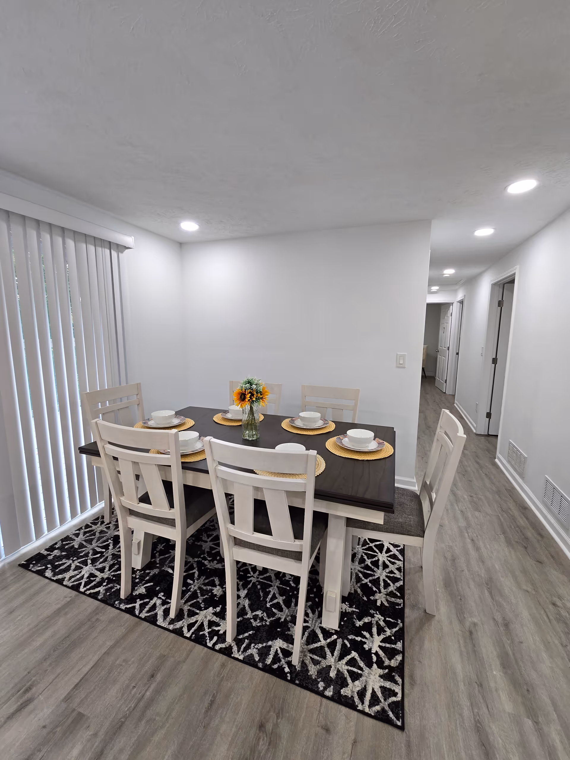Dining room with dark table, white chairs, sunflower centerpiece, and vertical blinds.