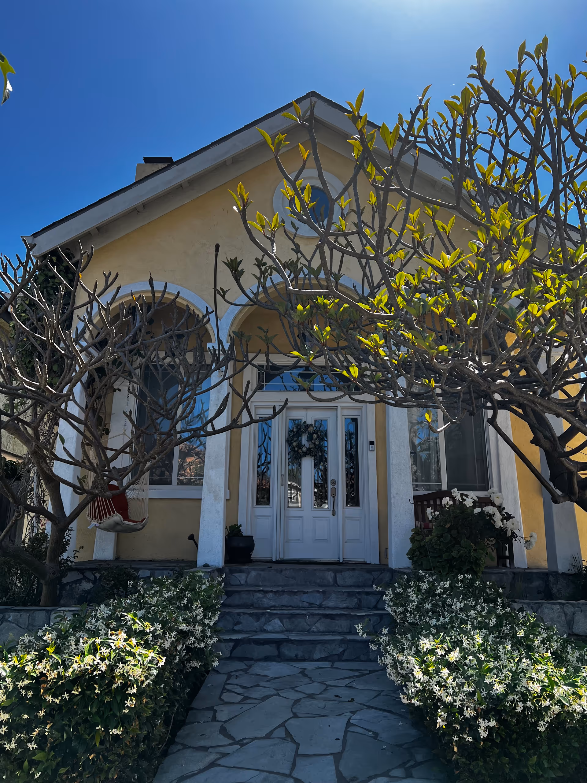Front entrance of a yellow house-style building framed by trees and flowering shrubs.