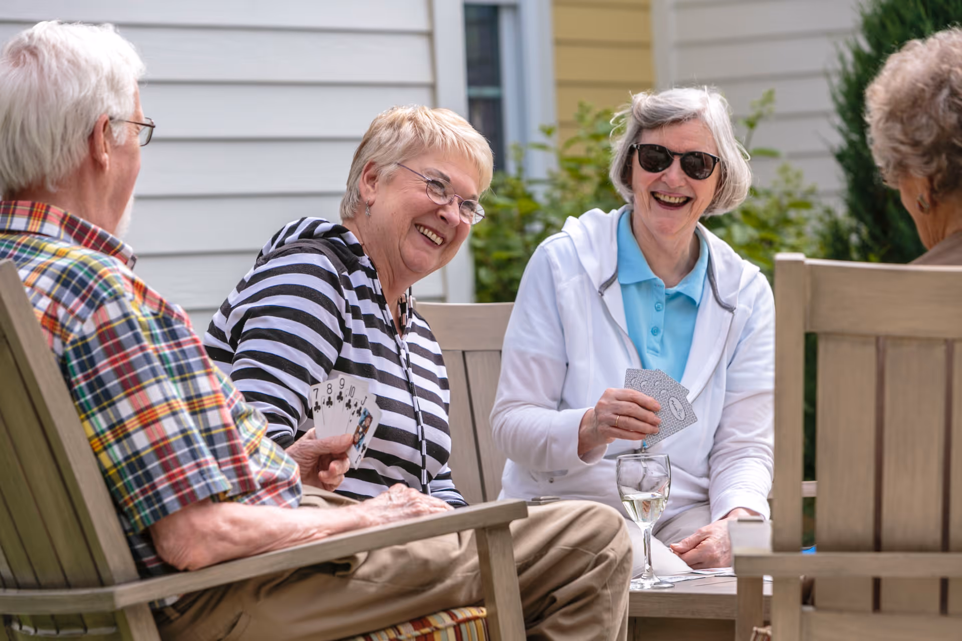 Four elderly people sitting outdoors on wooden chairs around a table, playing cards and smiling. One woman is wearing sunglasses and holding cards, another woman in a striped hoodie is also holding cards, and a man in a plaid shirt is partially visible. There is a glass of white wine on the table.