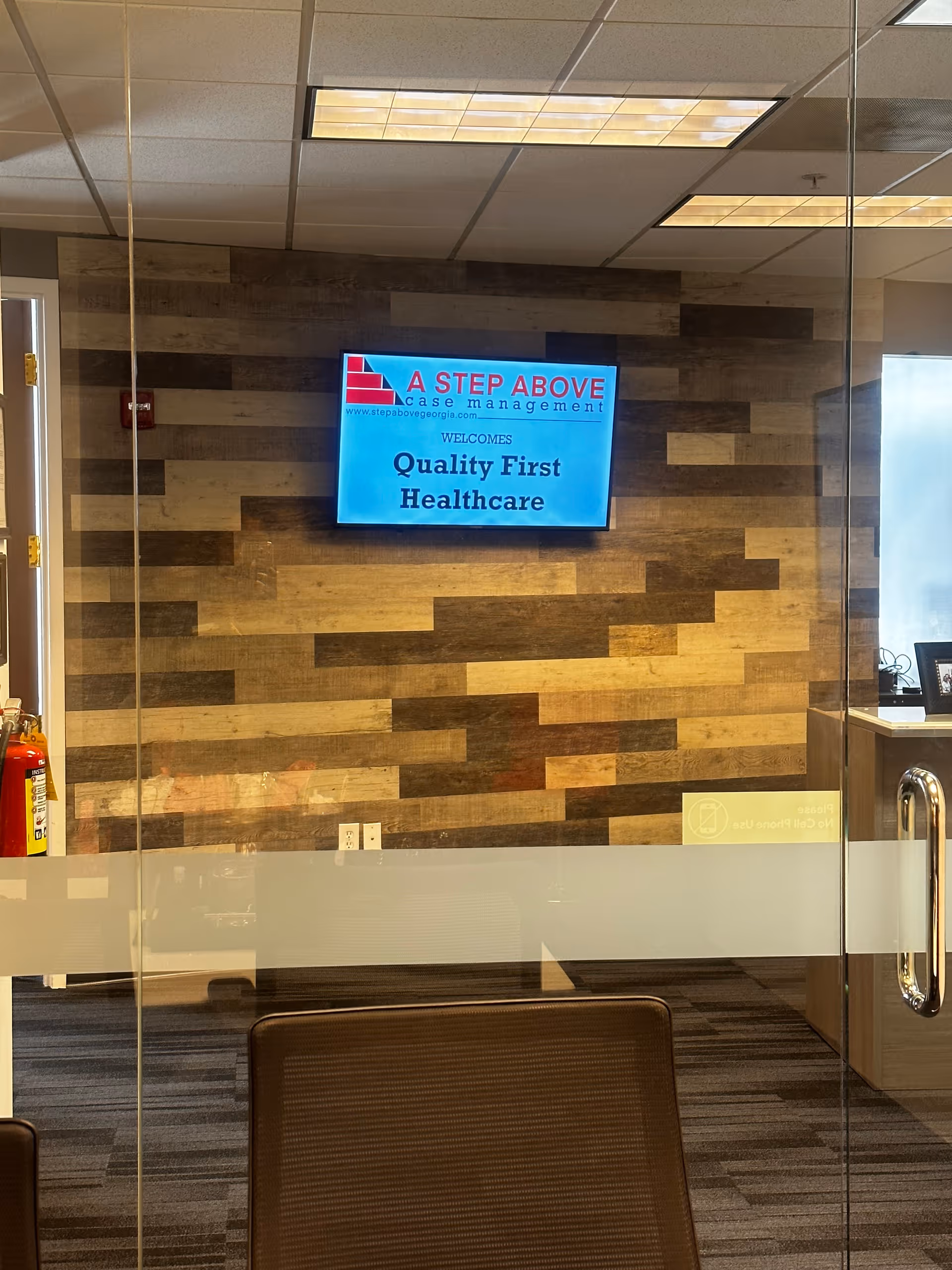 Interior reception area with wood-paneled wall, mounted screen, glass door, and brown chairs.