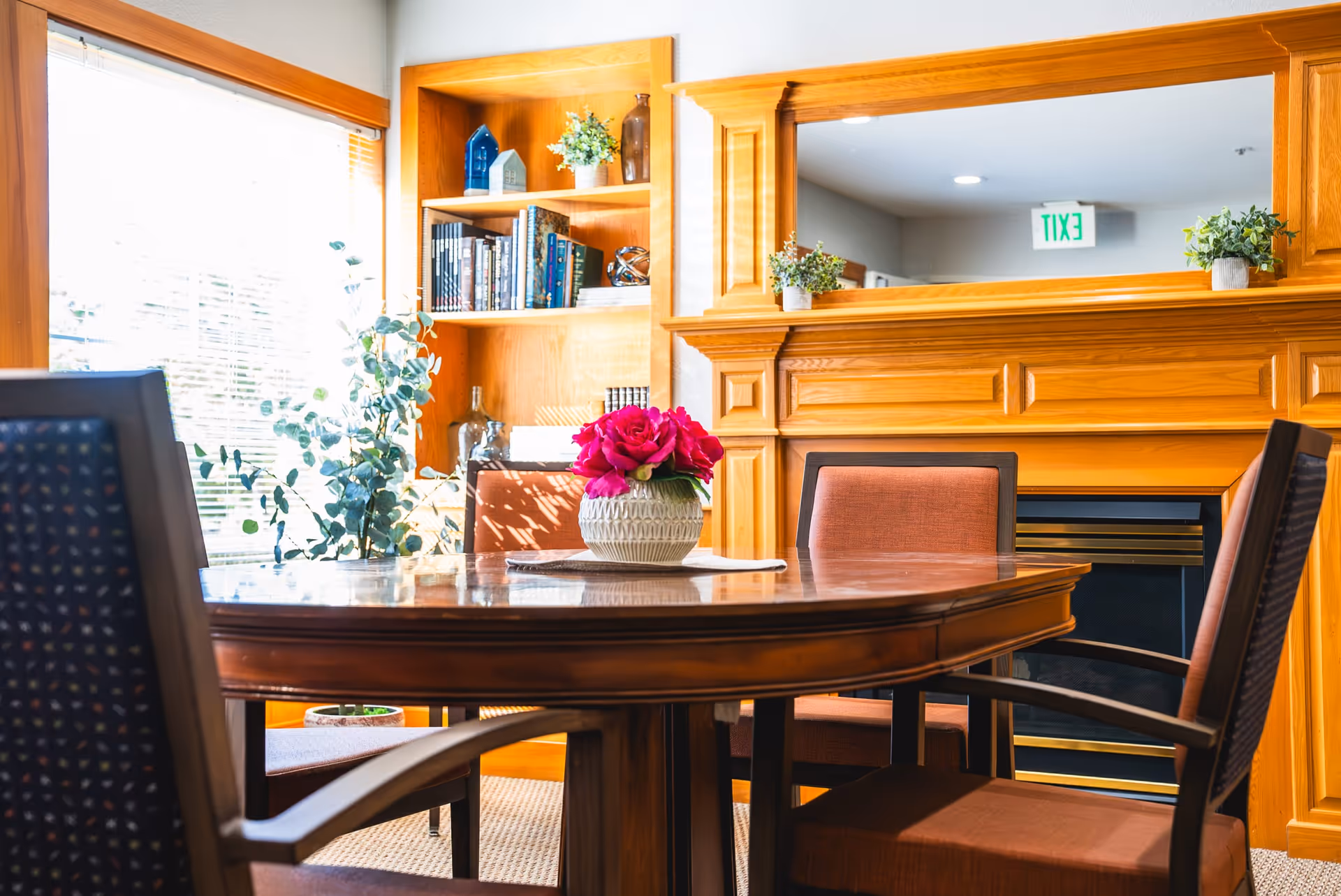 Round wooden dining table with chairs and a pink floral centerpiece in a bright room with built-in wooden shelving and a fireplace.