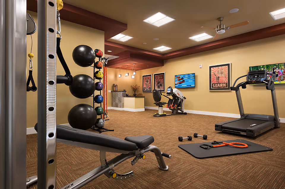 Interior of a senior living gym featuring treadmills, weights, stability balls, mats, and TVs.