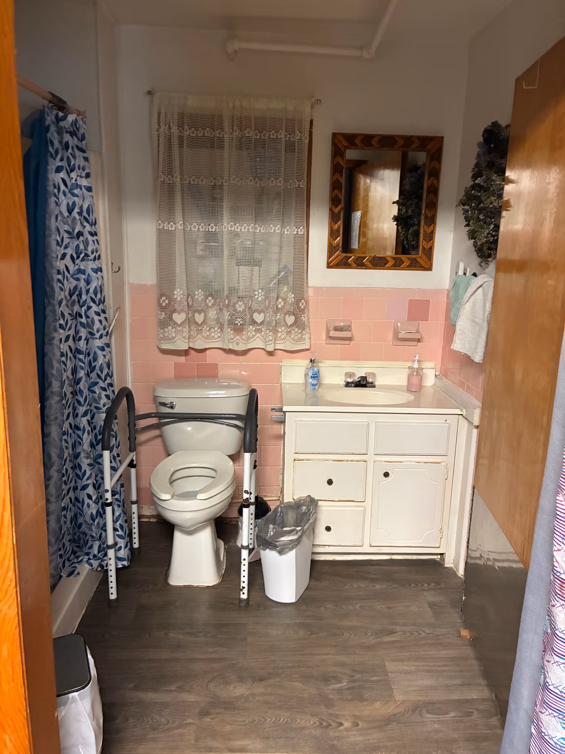 Accessible bathroom with pink tiles, toilet, walker, sink, and lace curtain.