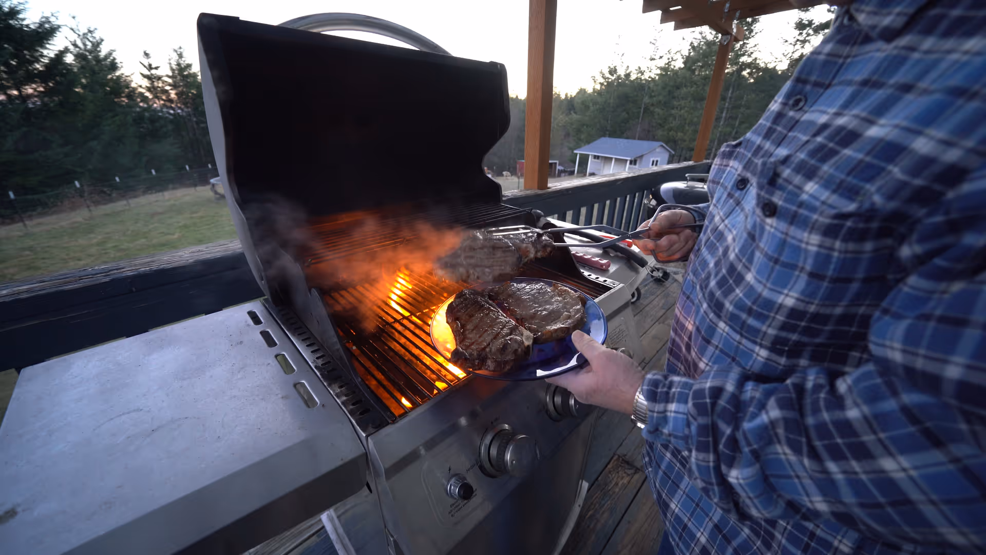 Person in plaid shirt grills steaks on a barbecue on an outdoor deck.