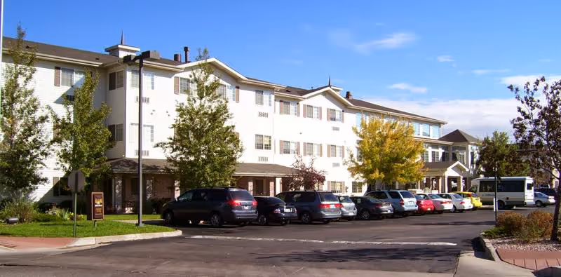 Front exterior of a white, multi-story senior living facility with parked cars and trees.