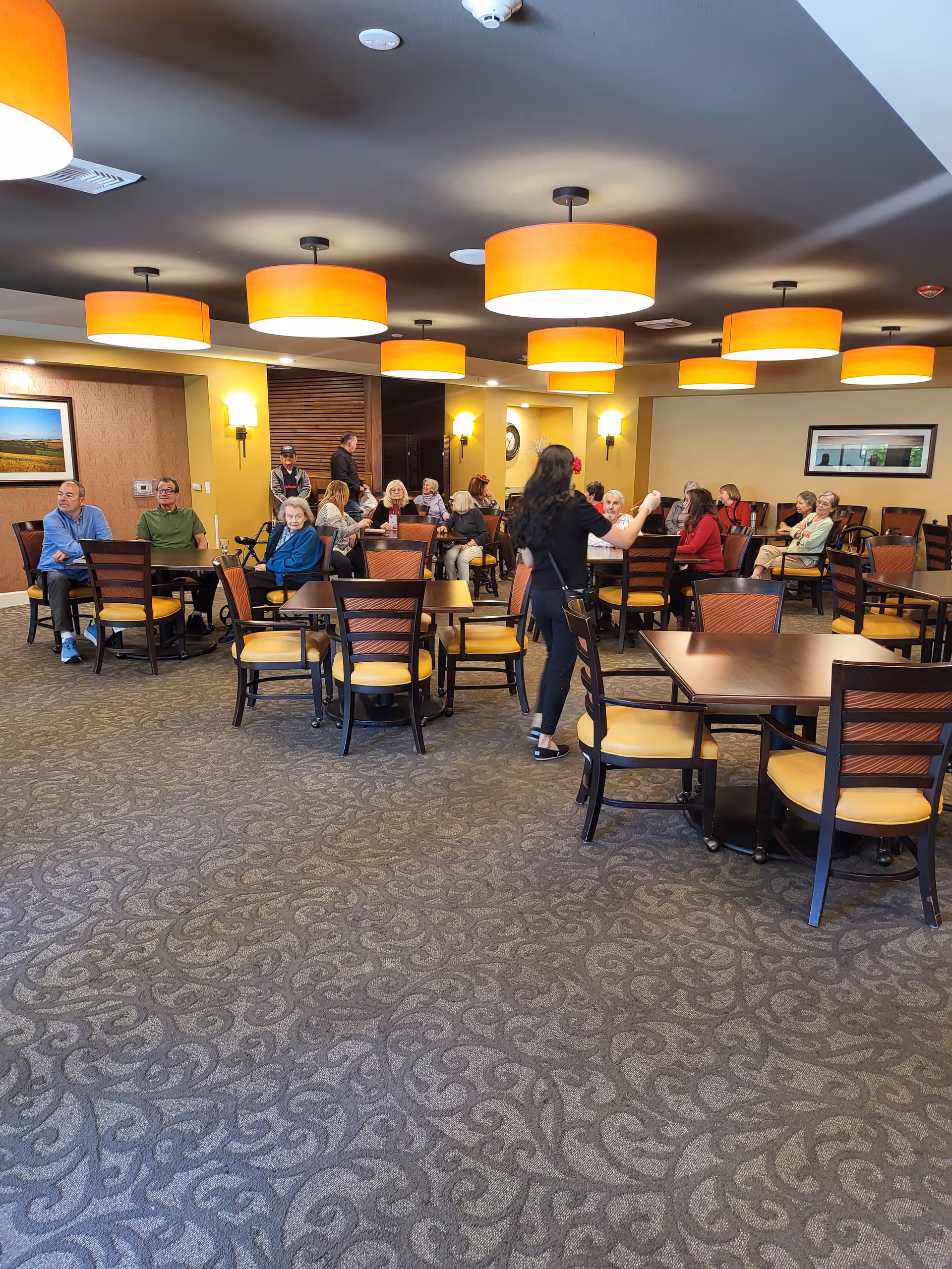 Senior residents and staff socialize around tables in a bright, spacious dining room.