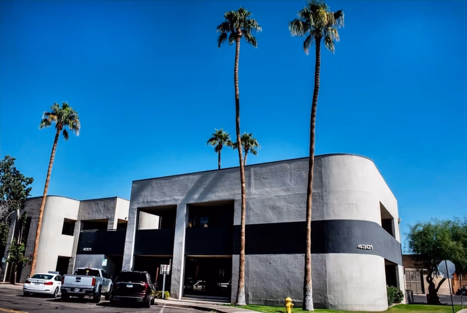 Modern light gray building with black stripe, palm trees, and parked cars in a sunny lot.
