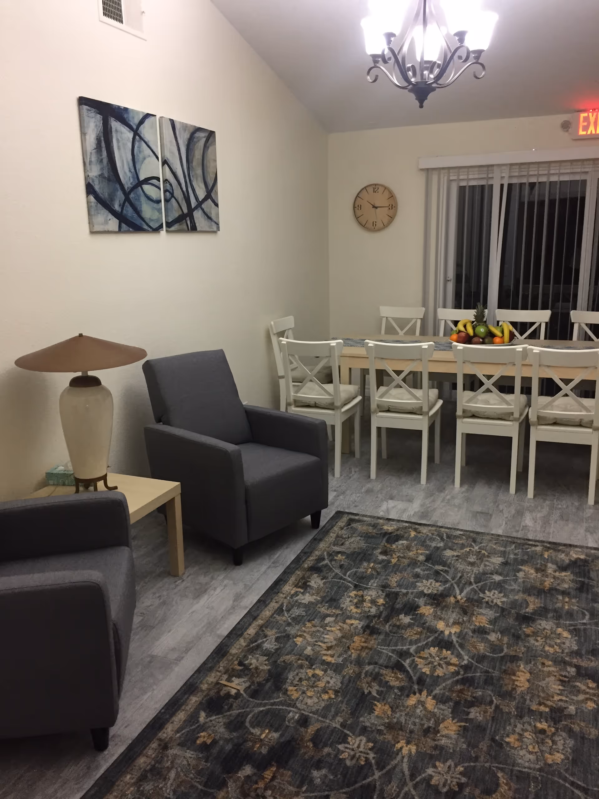 A bright dining area with a long table, white chairs, fruit bowl, clock, chandelier, and seating.