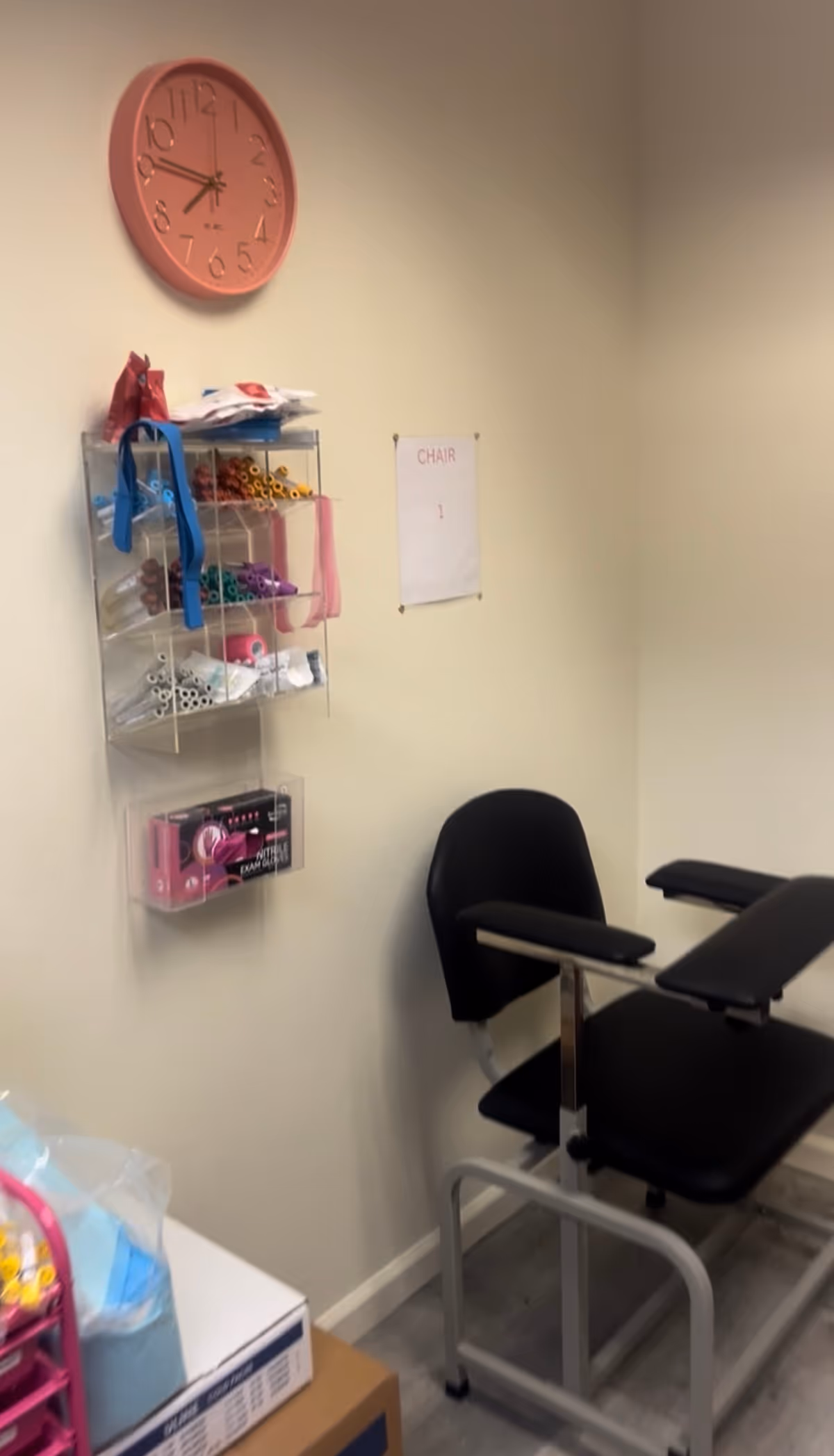 Pink clock above a transparent supply rack beside a black medical chair.