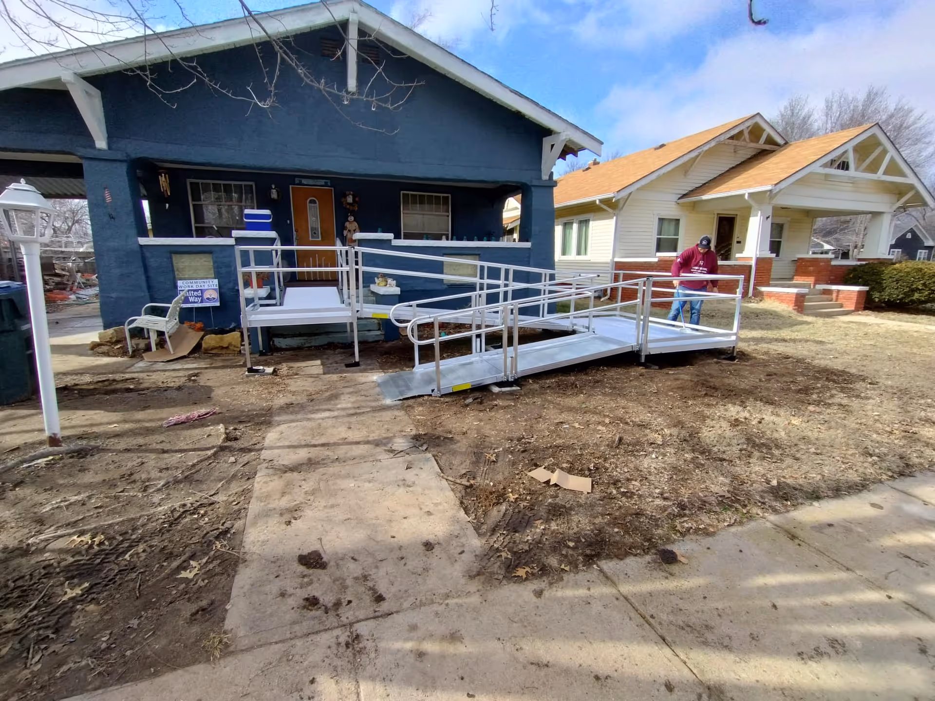 Wheelchair ramp extends to the front entrance of a blue house, with a person nearby.