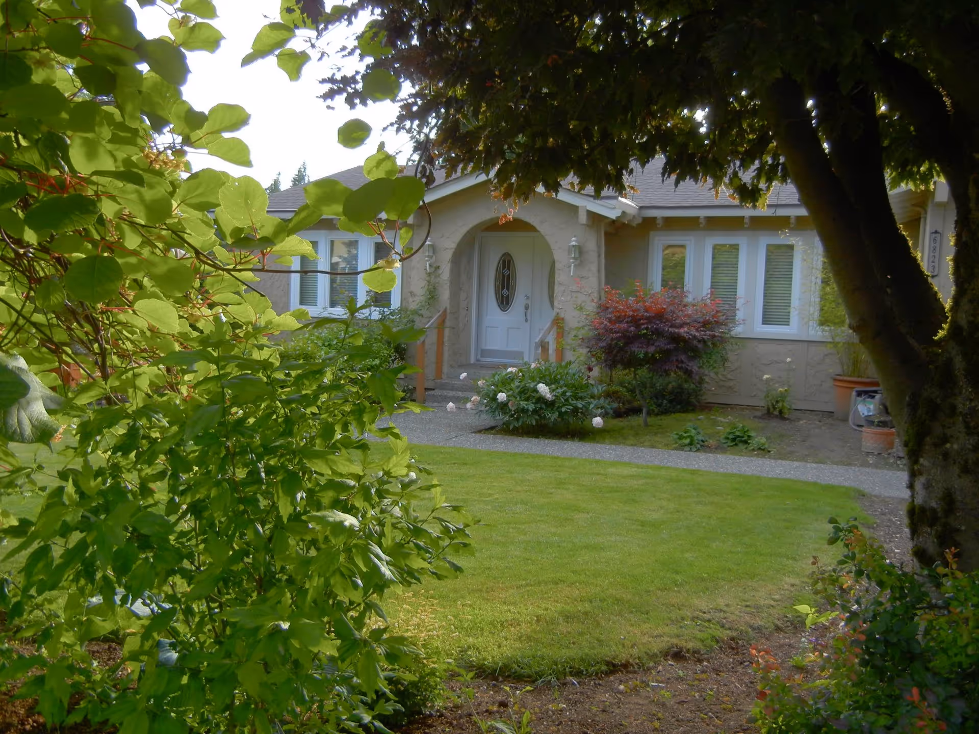 Front exterior of a beige house with curved entry, surrounded by lush greenery.