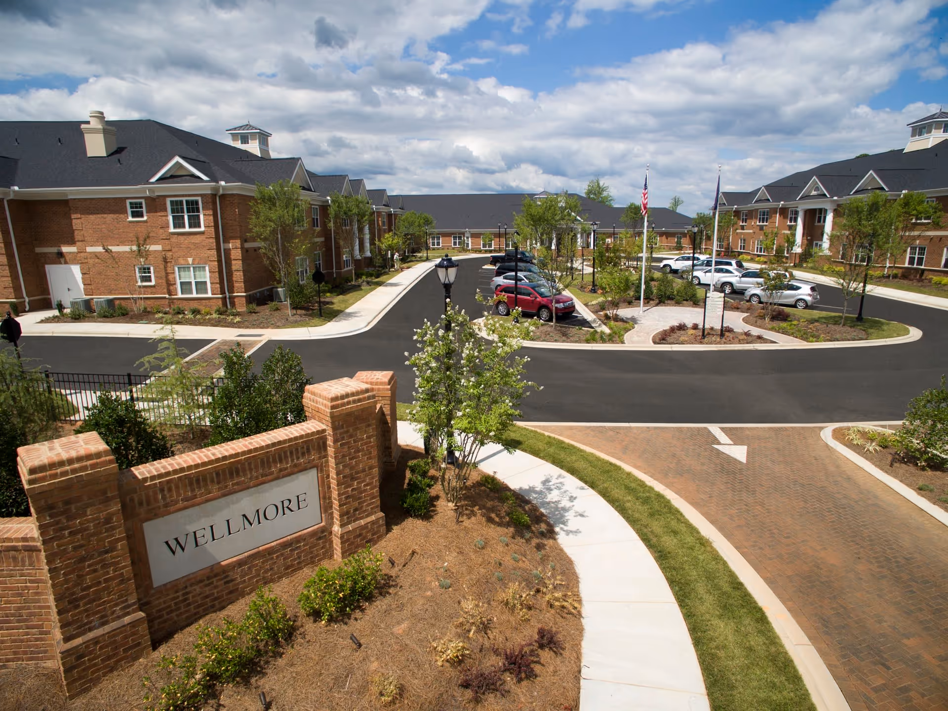 Entrance courtyard and parking area of a Wellmore senior living campus with brick buildings, landscaping, and a 'WELLMORE' entry sign.