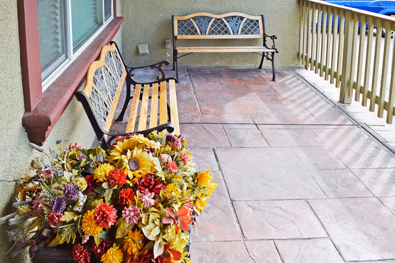 Outdoor patio area with two wooden benches featuring decorative metal backs, a large colorful flower arrangement in the foreground, a window with a red frame on the left wall, and a wooden railing on the right side.