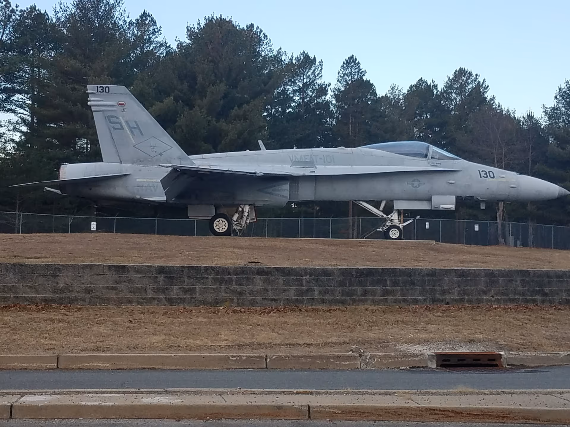 A gray fighter jet on outdoor display beside a fence and trees.