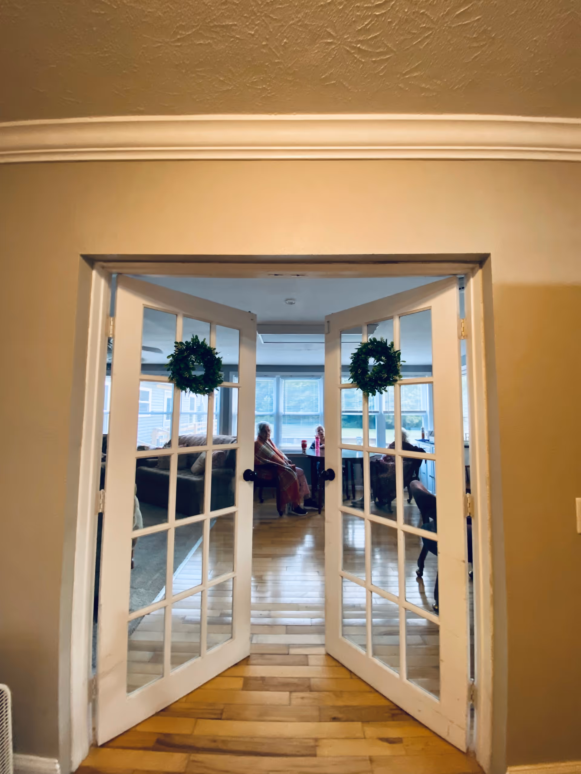 Open glass‑paneled double doors leading into a sunlit living room where residents sit by the windows.