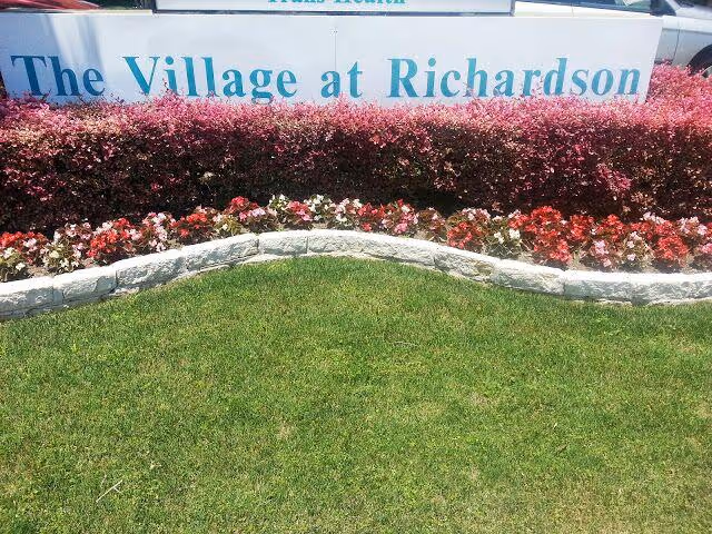 Sign reading The Village at Richardson with pink hedges and green lawn in front.