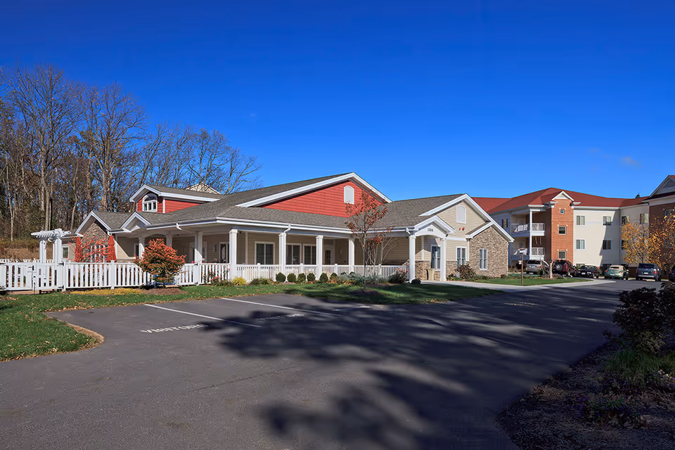Exterior view of a senior living facility building with beige and red siding, white trim, and a white picket fence. There is a parking lot in front with several cars parked and trees without leaves in the background under a clear blue sky.