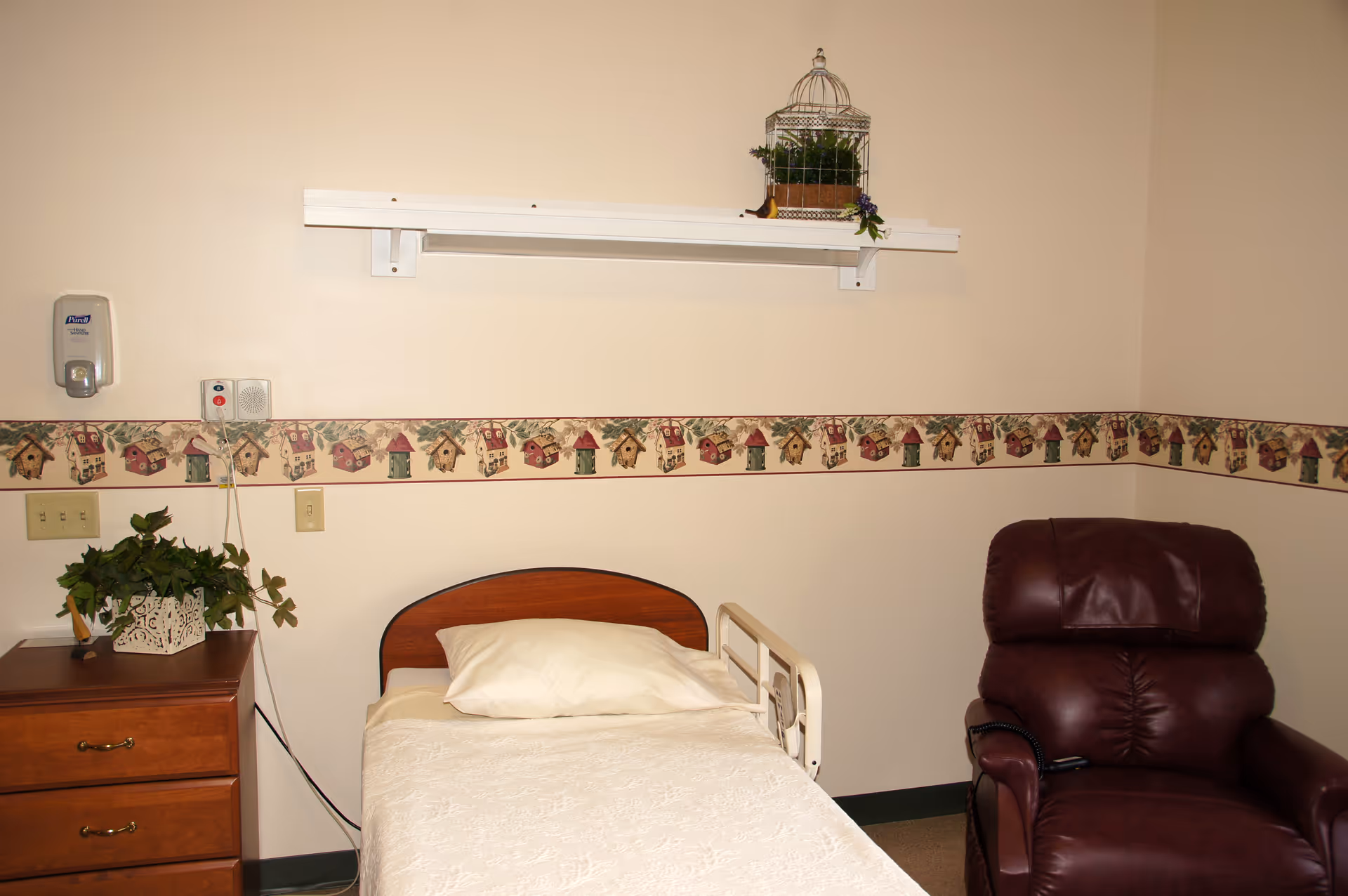 A small room with a single bed featuring a wooden headboard and white bedding, a brown leather recliner chair, a wooden nightstand with a potted plant, and a decorative wallpaper border with birdhouses along the wall.