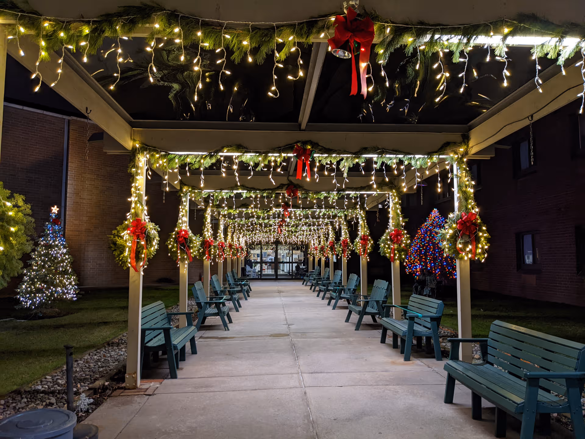 Outdoor covered walkway illuminated by festive lights with wreaths and teal benches at night.