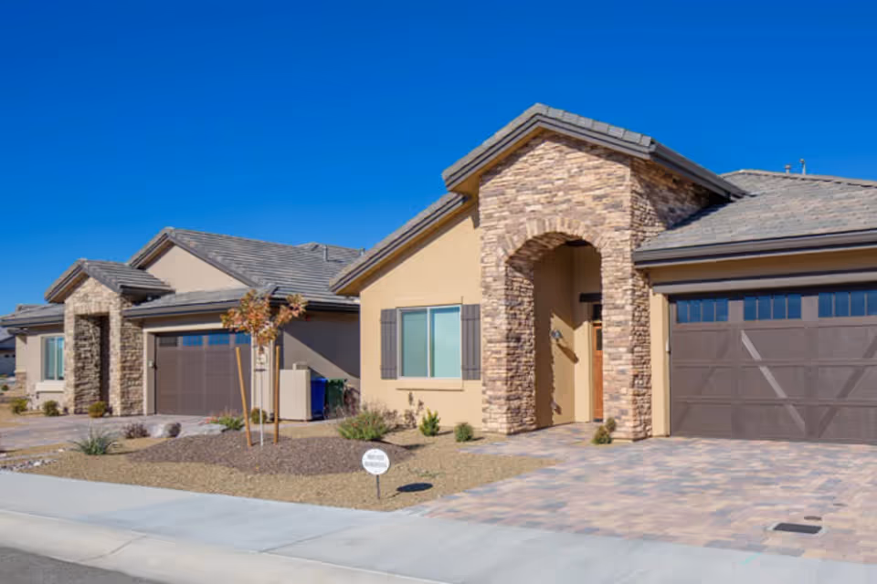 Exterior view of a single-story residential building with stone and stucco facade, featuring a garage with a brown door, a small front yard with desert landscaping, and a clear blue sky.