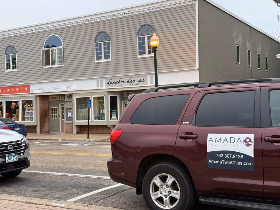 A maroon Amada Senior Care van parked in front of storefronts along the street.