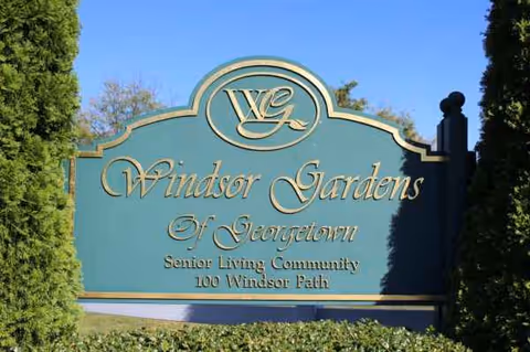 Teal Windsor Gardens sign at the senior living facility entrance, with surrounding trees.