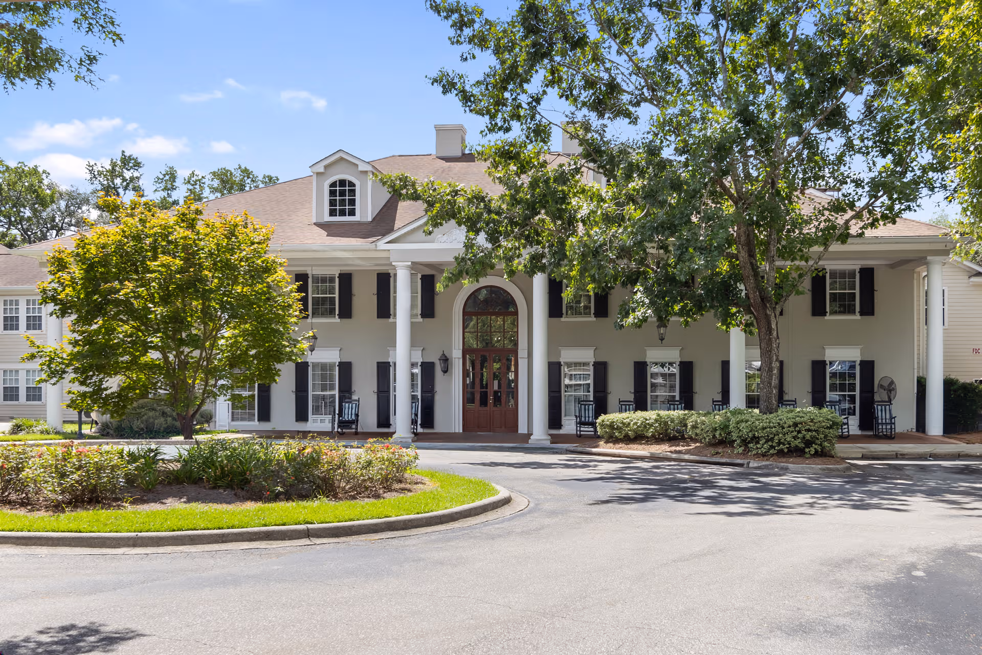 Beige senior living facility exterior with columns, trees, and a central arched doorway.