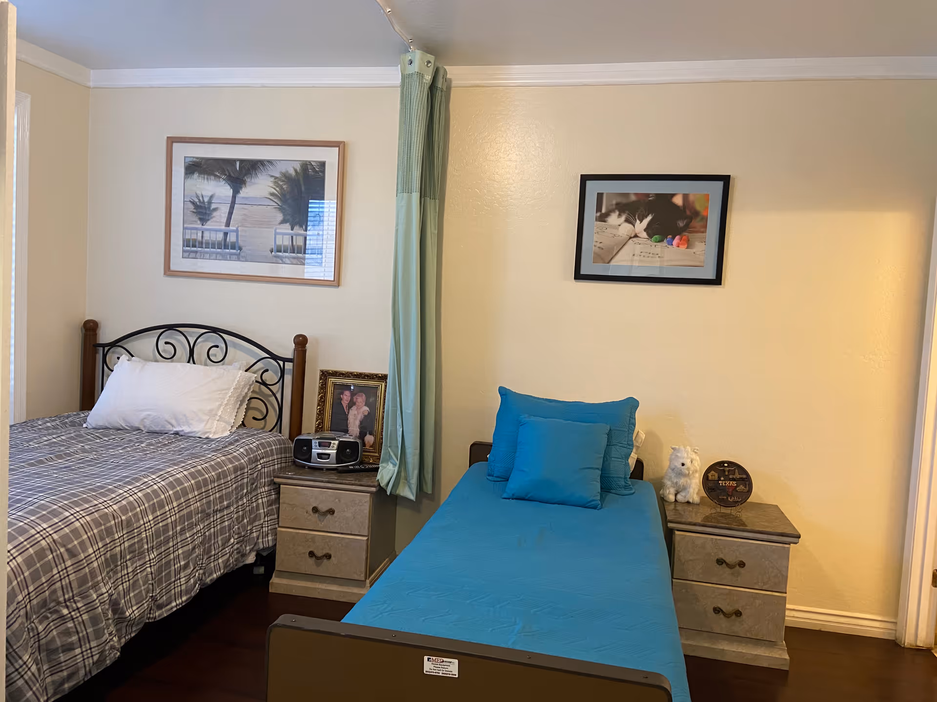 Two beds in a beige bedroom with a nightstand, framed photos, and blue cushions.