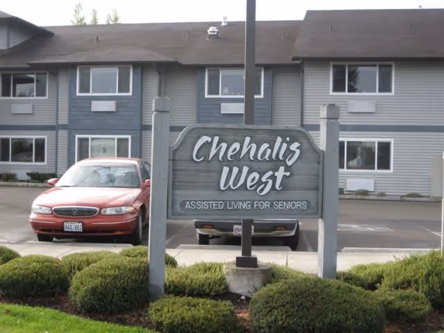 Entrance sign reading 'Chehalis West Assisted Living for Seniors' with landscaped bushes, parked cars, and the two-story facility building behind it.