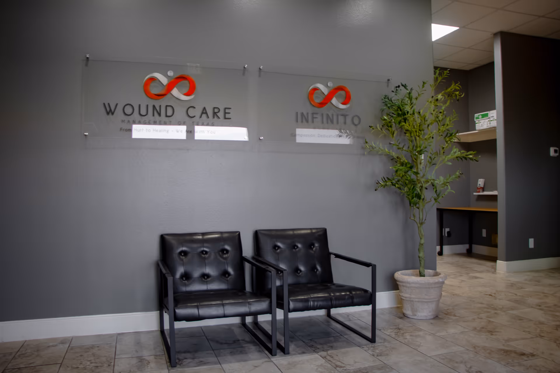 Lobby waiting area with two black chairs, wall signage for Wound Care and Infinito, potted plant.