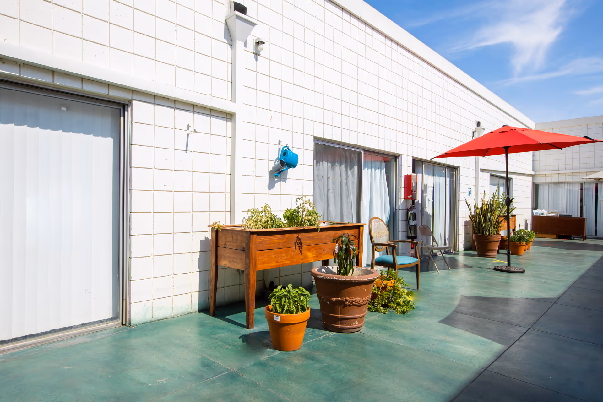 Outdoor patio area with potted plants, a wooden planter box, chairs, and a red umbrella attached to a stand. The patio is adjacent to a white building with large windows and sliding doors under a clear blue sky.