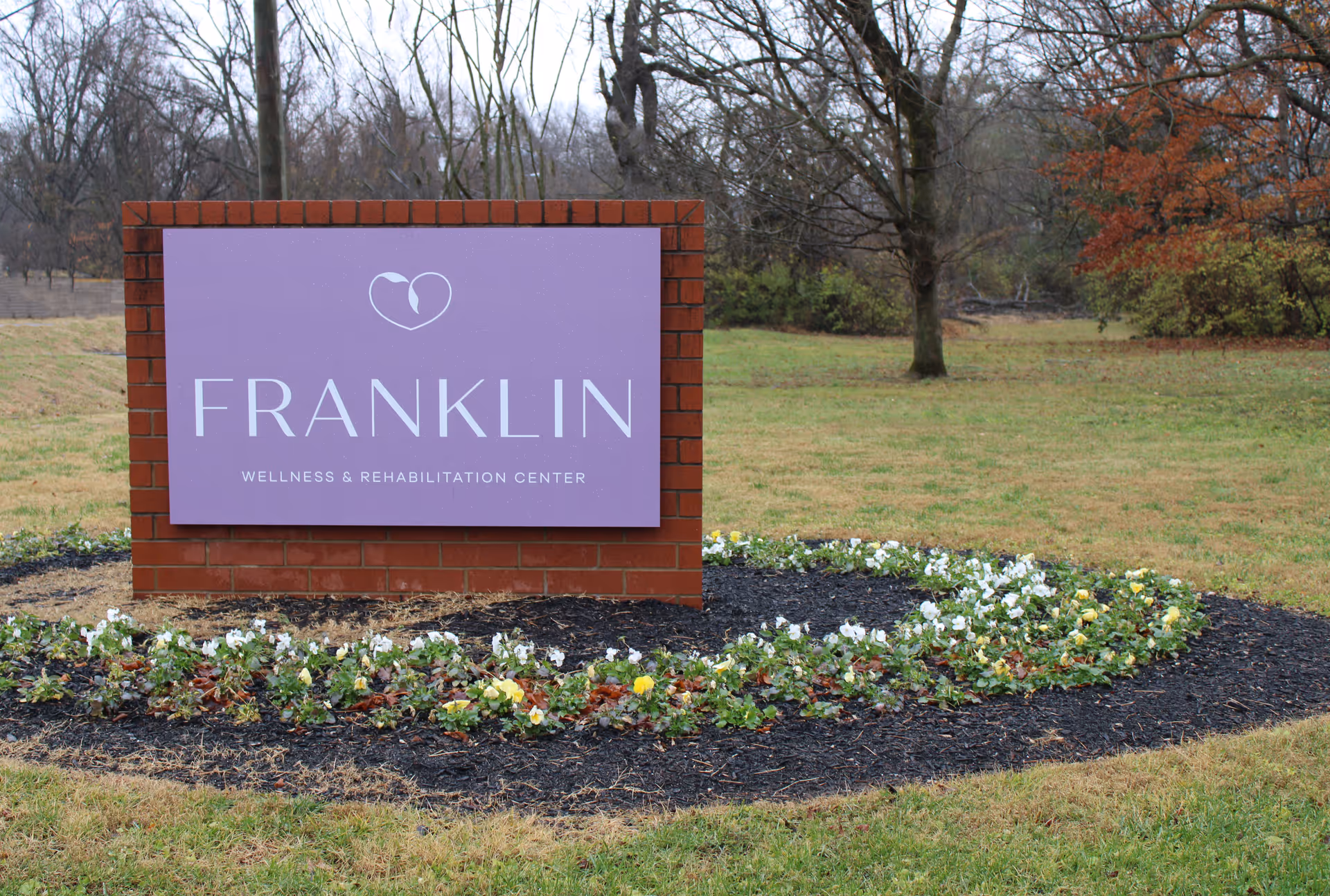Purple Franklin sign on a brick pedestal in a landscaped flower bed outdoors.