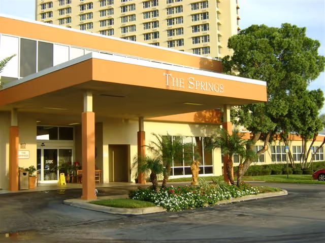 Front entrance of a senior living facility with an orange canopy, palm trees, and landscaping.