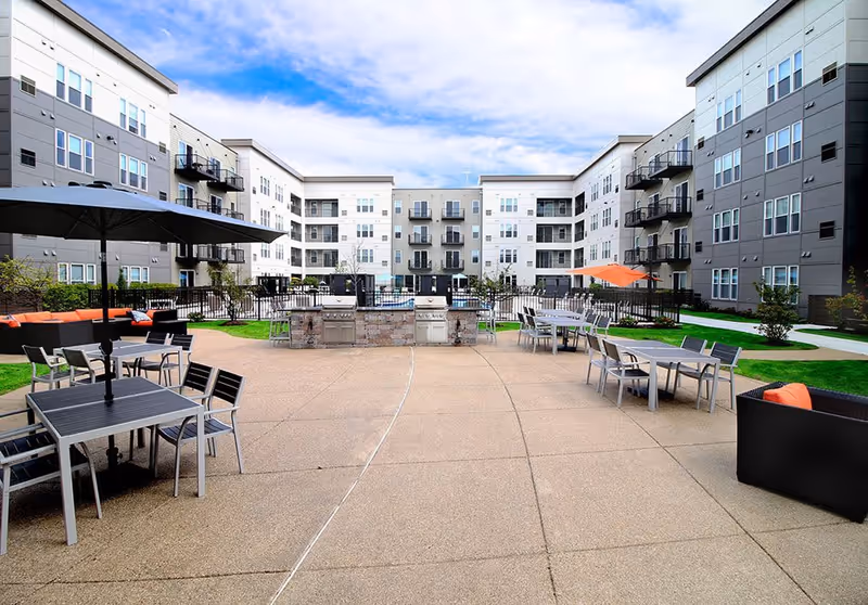 Outdoor courtyard of a multi-story residential building with patio tables, umbrellas, lounge seating, grills and a fenced pool area.