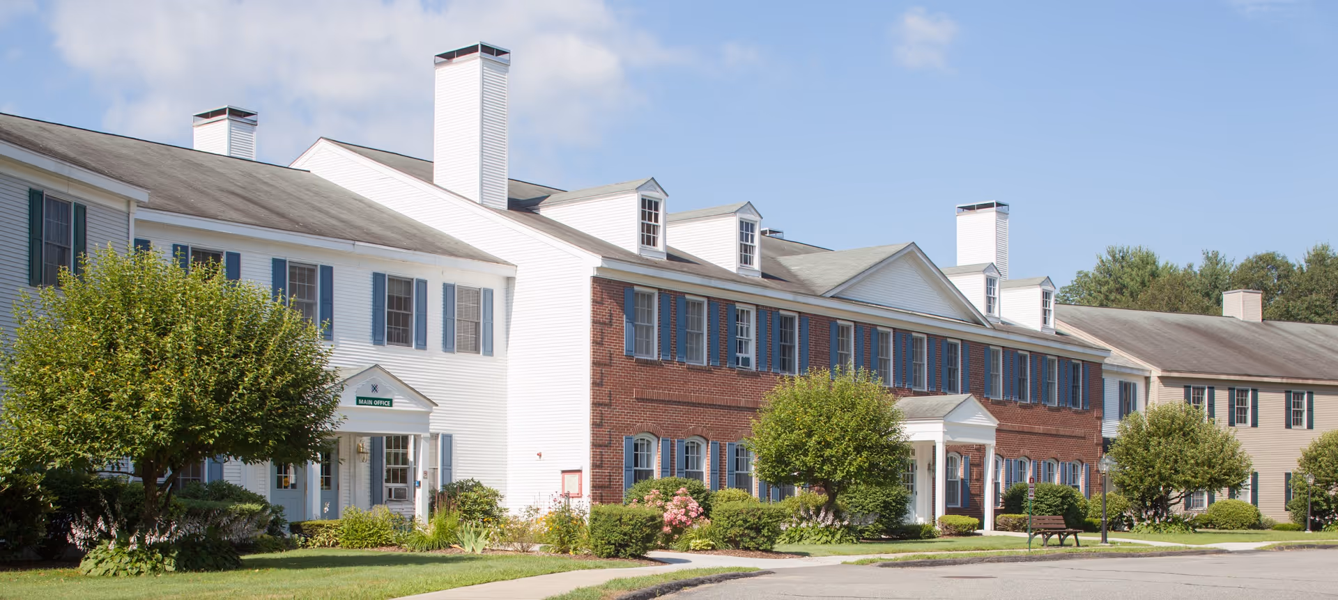 Front exterior of a two-story senior living facility with brick and white siding, landscaped lawns, and a sidewalk.
