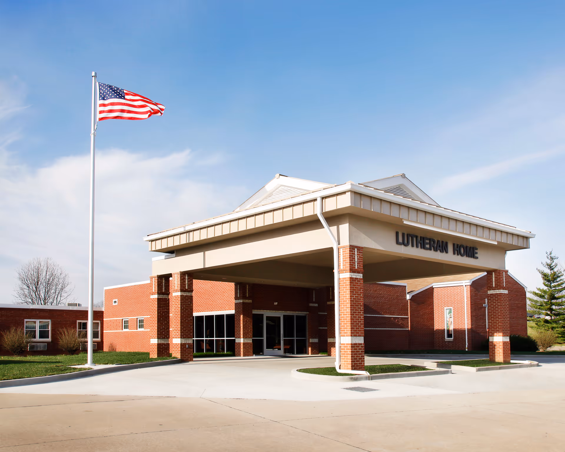 Sunny day view of Lutheran Home exterior with brick entrance, porte-cochère, and American flag.