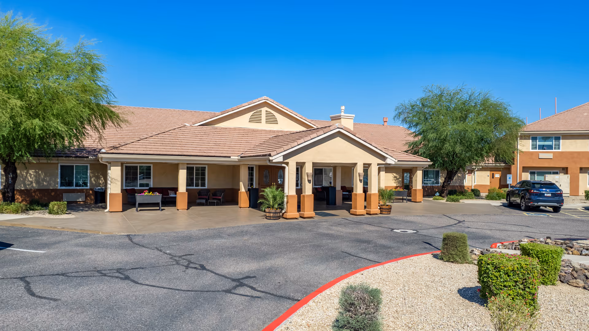 Front entrance of a beige single-story senior living building with a covered portico, driveway, desert landscaping and a parked car.