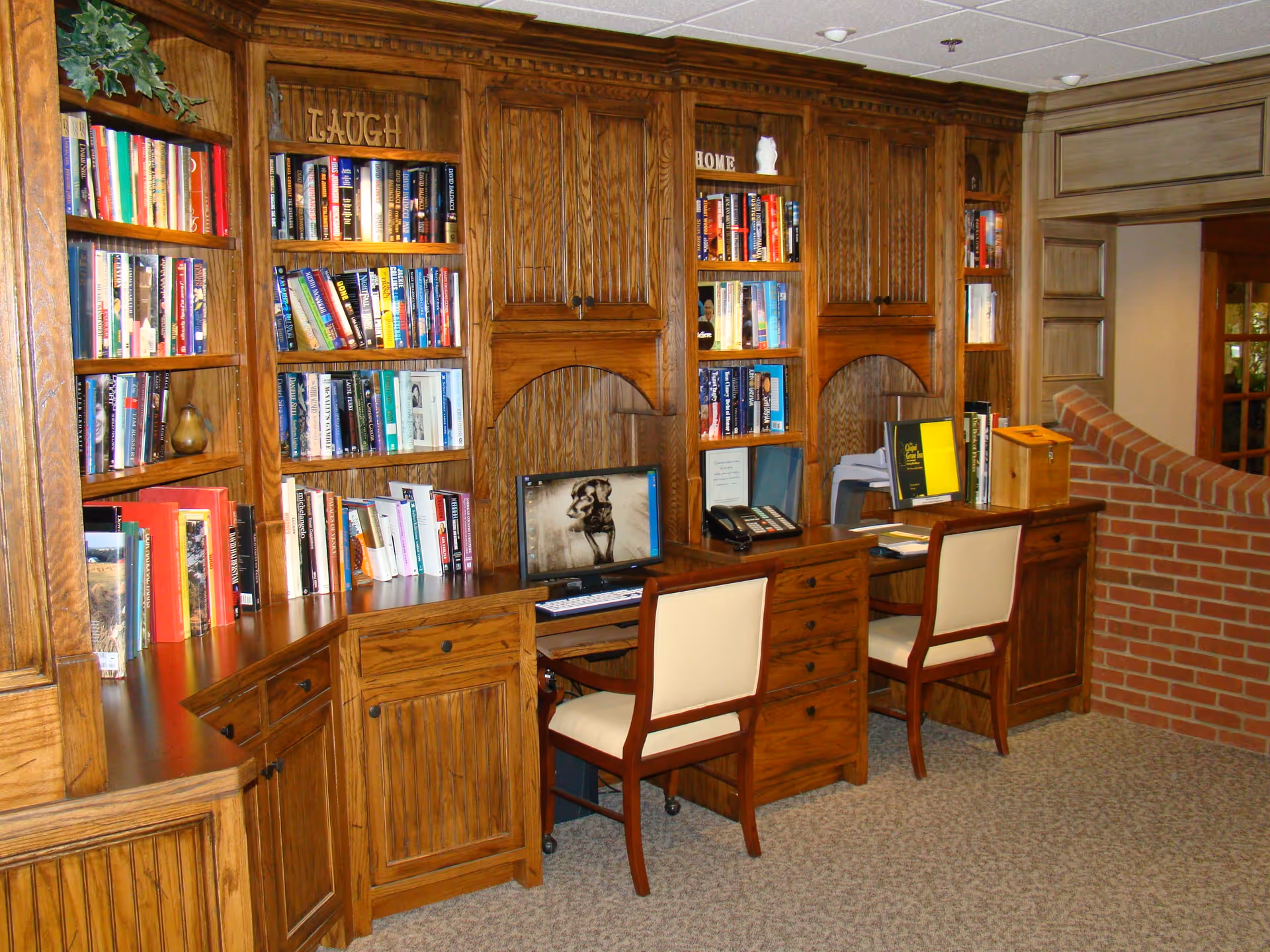A warm wood-paneled library/office area with built-in bookshelves, desks, chairs, and a computer.