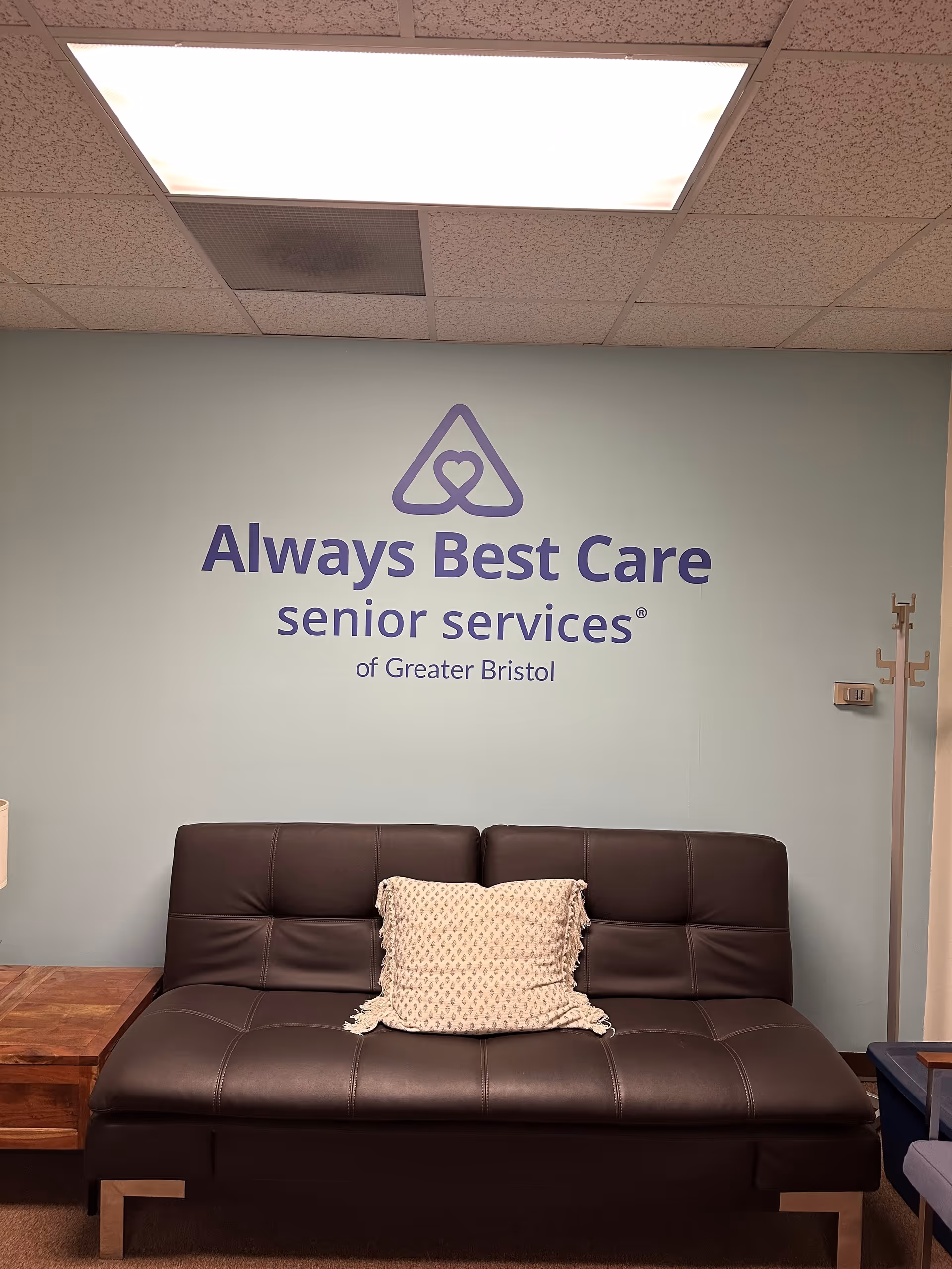 Lobby seating area with a brown sofa and Always Best Care logo on pale teal wall.