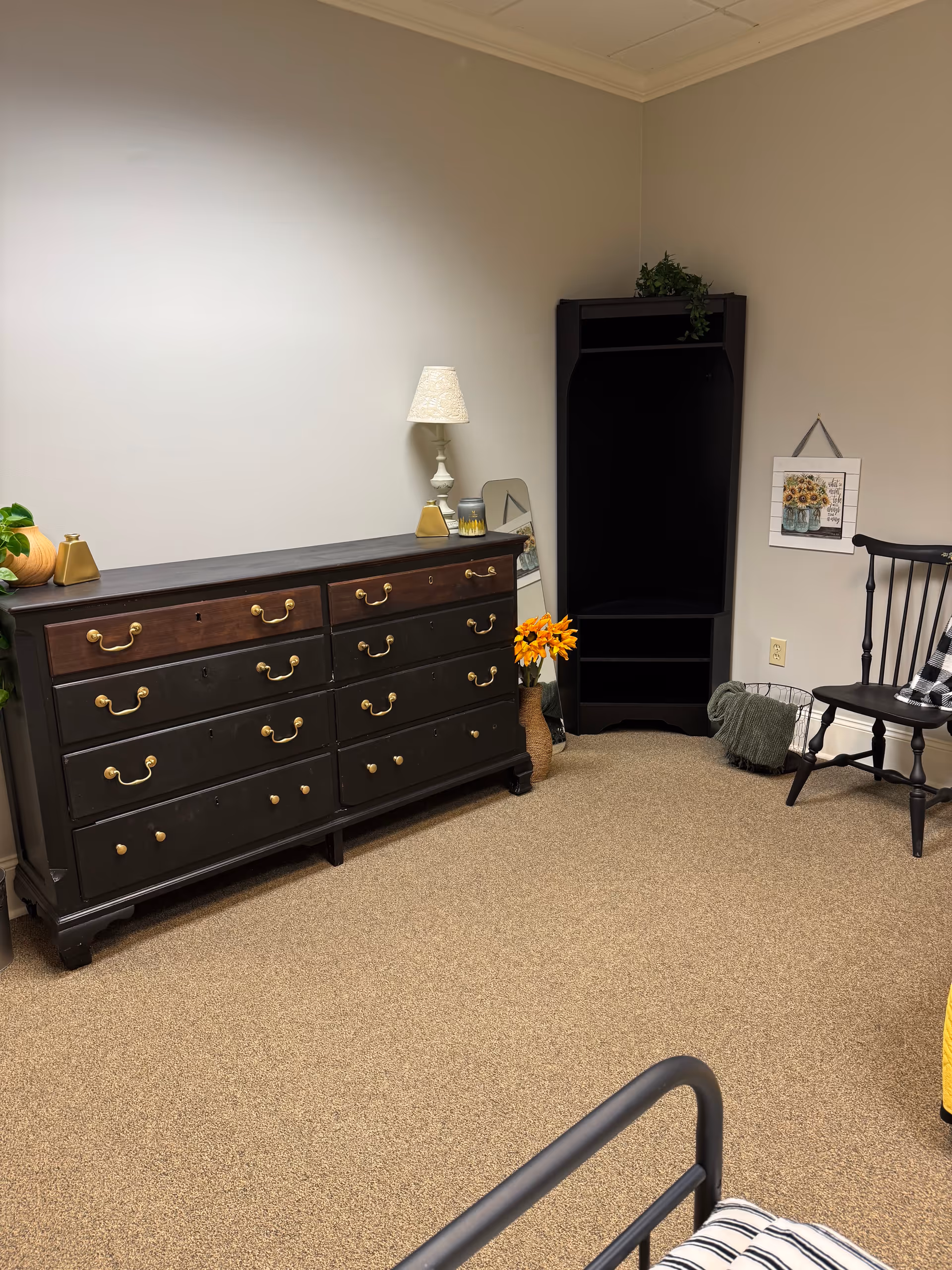 A cozy, sparsely furnished bedroom with a long dresser, lamp, black armoire, chair, and wall art.