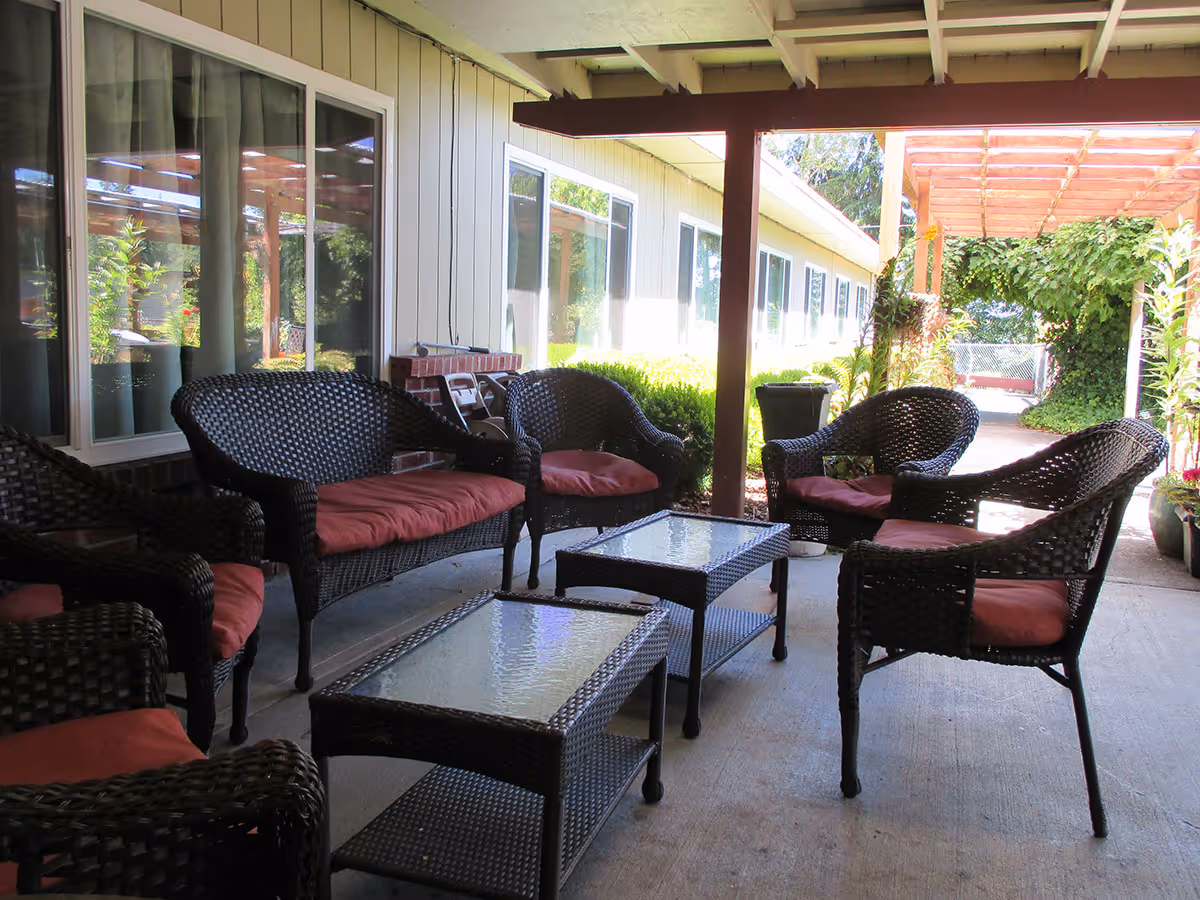 Outdoor covered patio area with wicker chairs and sofas with red cushions arranged around glass-top coffee tables, adjacent to a building with large windows and surrounded by greenery.
