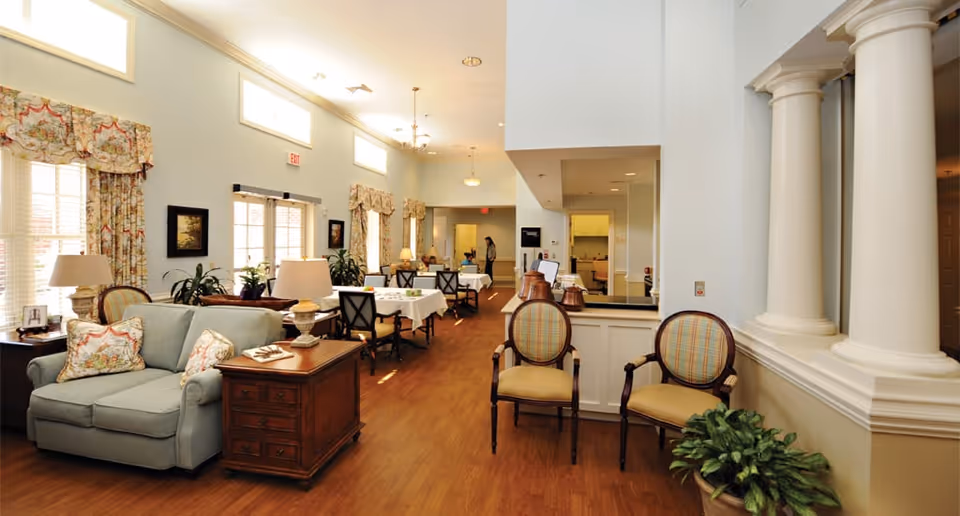 A bright and spacious interior room with wooden flooring, featuring a light blue upholstered loveseat with floral cushions, a wooden side table with a lamp, and several dining tables with chairs covered in white tablecloths. The room has large windows with floral curtains, potted plants, and decorative columns on the right side.