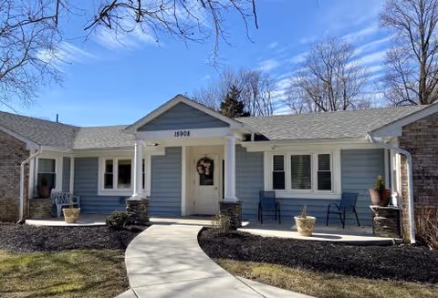 A single-story care home exterior with a curved walkway, front porch, and blue chairs.